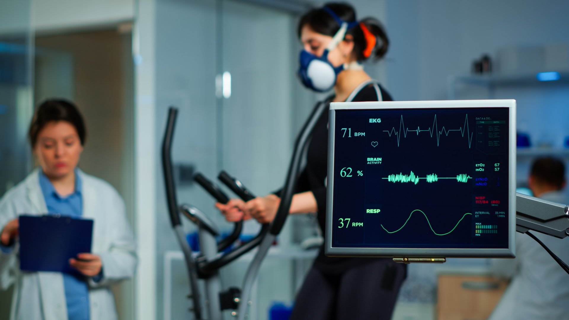 Monitor showing EKG scan of athlete running on cross trainer and medical specialists supervise exercise in background, controlling physical activity, measuring heart rate in sports science laboratory